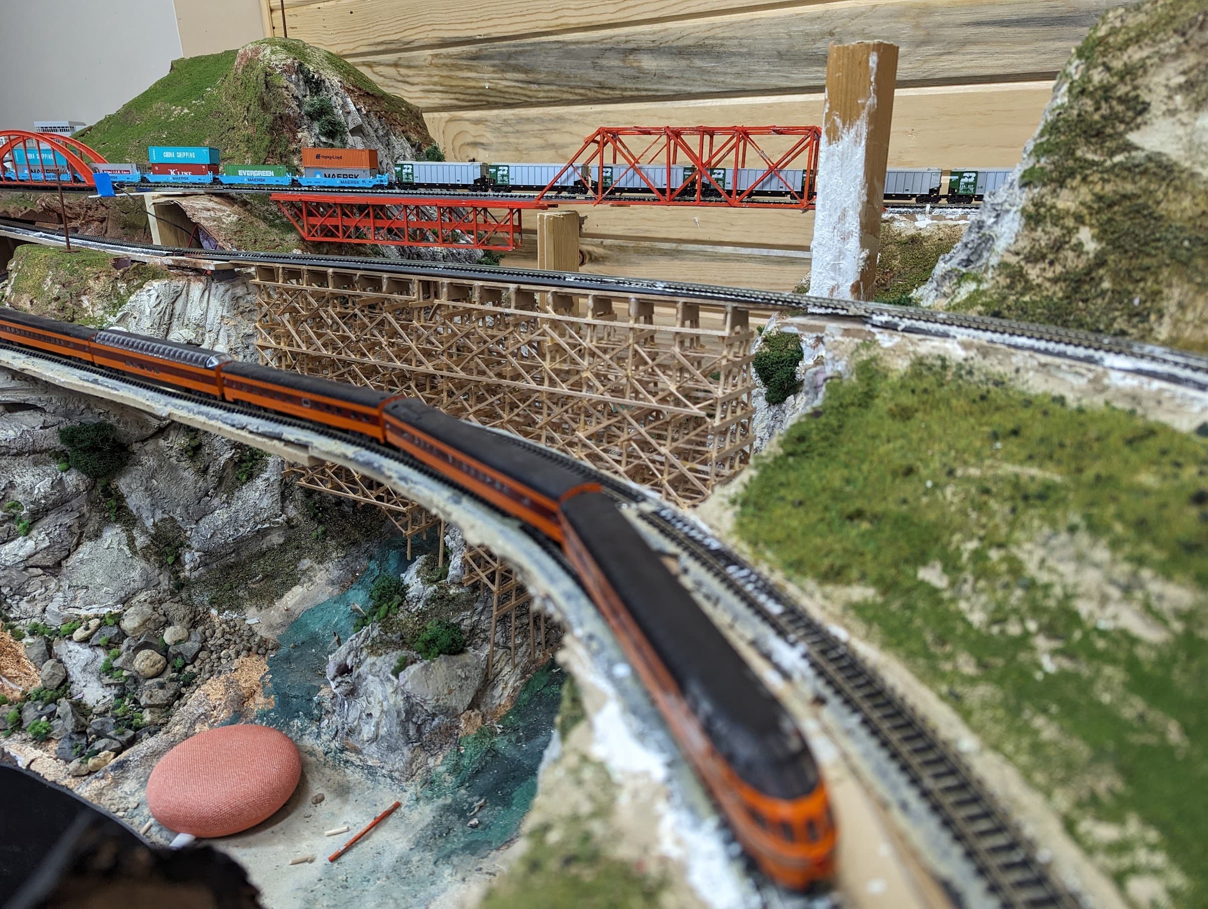 A passenger train rounding a curve below a wooden trestle, with a freight crossing a red truss bridge behind it